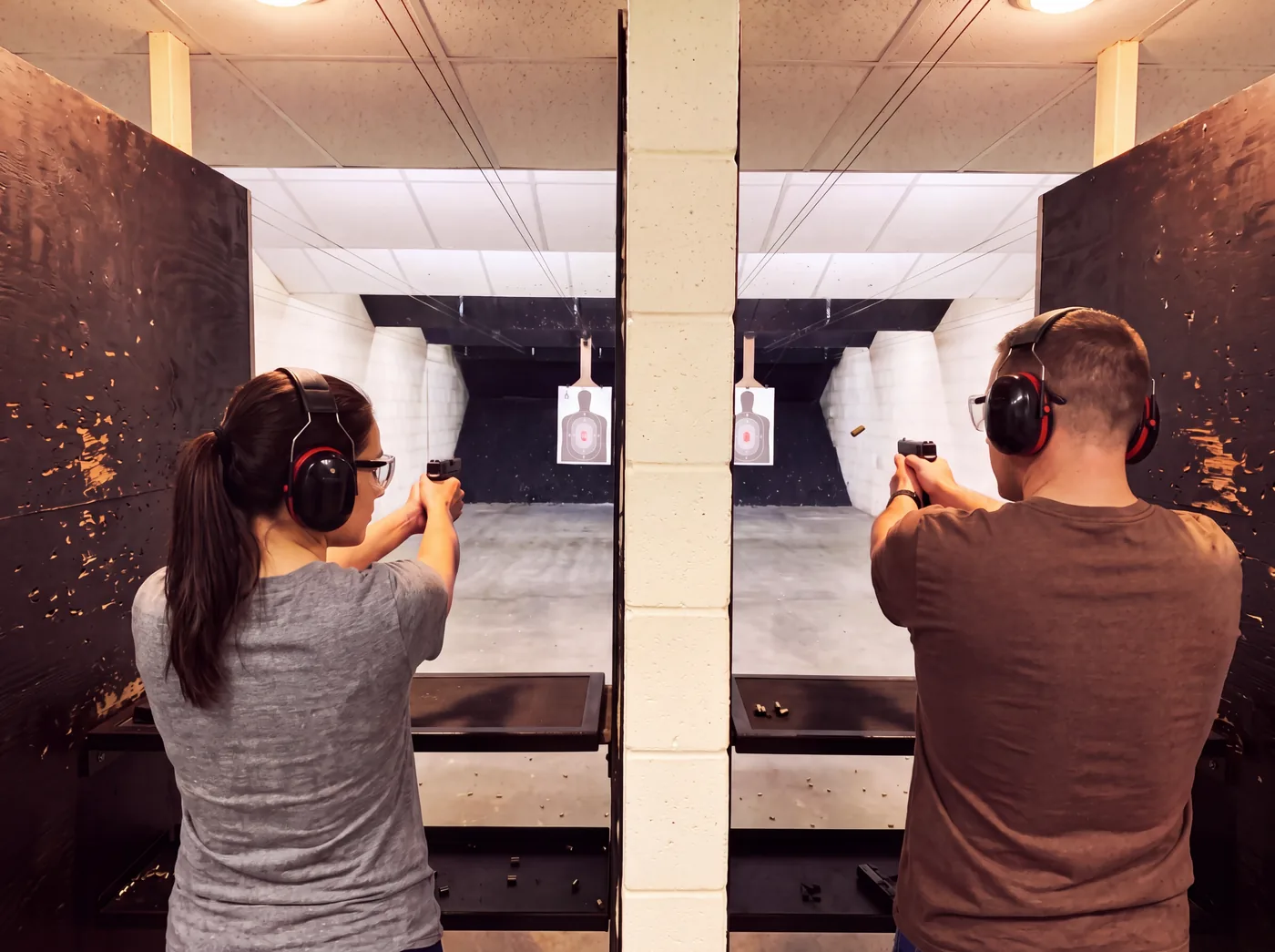 Two friends in adjacent lanes on the indoor range, ear and eye protection on, each shooting at their own target.
